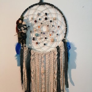 Può includere: Un acchiappasogni bianco e nero con piume blu e bianche. L'acchiappasogni è decorato con perline e una piccola bambola.