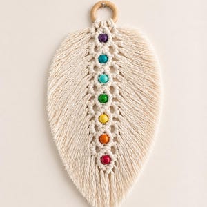 Puede incluir: Colgante de pared de macramé color crema en forma de hoja, adornado con una serie de cuentas coloridas. Las cuentas están dispuestas verticalmente, mostrando un espectro de colores. Un anillo de madera está en la parte superior para colgar.