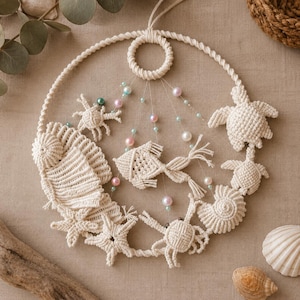 Pode incluir: Uma decoração de parede circular em macramé com formas de corda atada em branco sujo que representam um caranguejo, uma estrela-do-mar, um peixe e tartarugas marinhas. O design é acentuado com pequenas contas iridescentes em tons rosa, azul e branco.