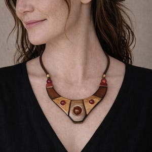 Può includere: Collana in legno dal design geometrico. La collana presenta un cordino in pelle marrone, elementi in legno e dettagli di pietre preziose rosse. La donna nell'immagine indossa un top nero.