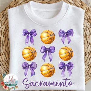 May include: White sweatshirt with a design of basketballs and purple bows. The word "Sacramento" is written in a script font below the design. The sweatshirt is displayed on a woven basket.