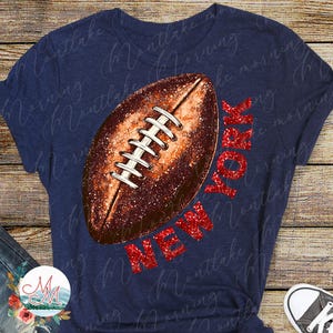 May include: Navy blue t-shirt with a football design and the text "NEW YORK" in red glitter. The football is brown with white laces. A casual wear item.