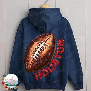 K&ouml;nnte beinhalten: Marineblauer Kapuzenpullover mit einem gro&szlig;en Football-Motiv und dem Wort "HOUSTON" in rotem Glitzer. Der Football ist braun mit wei&szlig;en Schn&uuml;rsenkeln. Der Kapuzenpullover hat eine Kapuze und einen Kordelzug.