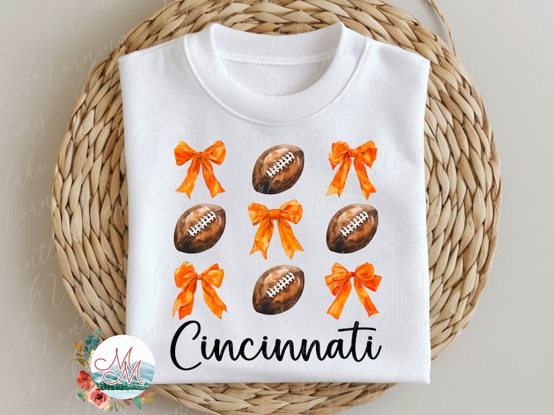 Peut inclure: Sweat-shirt blanc avec des illustrations &agrave; l'aquarelle de ballons de football am&eacute;ricain et de n&oelig;uds orange. Le mot "Cincinnati" est &eacute;crit en noir. Le motif est centr&eacute; sur le devant du v&ecirc;tement.