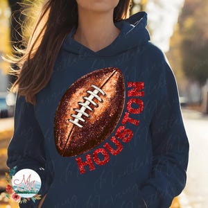 K&ouml;nnte beinhalten: Marineblauer Hoodie mit einem Football-Design und dem Wort "HOUSTON" in rotem Glitzer. Der Football ist braun mit wei&szlig;en Schn&uuml;rsenkeln. Ein l&auml;ssiges, sportliches Kleidungsst&uuml;ck.