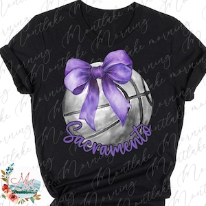 May include: Black t-shirt with a basketball design. The design features a purple bow, a basketball, and the word "Sacramento" in purple script. The design is centered on the shirt, making it a striking piece.