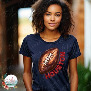 K&ouml;nnte beinhalten: Marineblaues T-Shirt mit einem Football-Motiv und dem Wort "HOUSTON" in rotem Glitzer. Der Football ist braun mit wei&szlig;en Schn&uuml;rsenkeln. Das T-Shirt hat einen Rundhalsausschnitt und kurze &Auml;rmel.