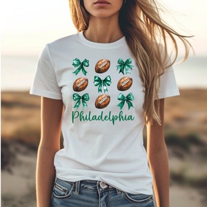 Op de afbeelding: Wit T-shirt met een groen en bruin voetbal- en strikdesign. De tekst "Philadelphia" is op het shirt gedrukt.