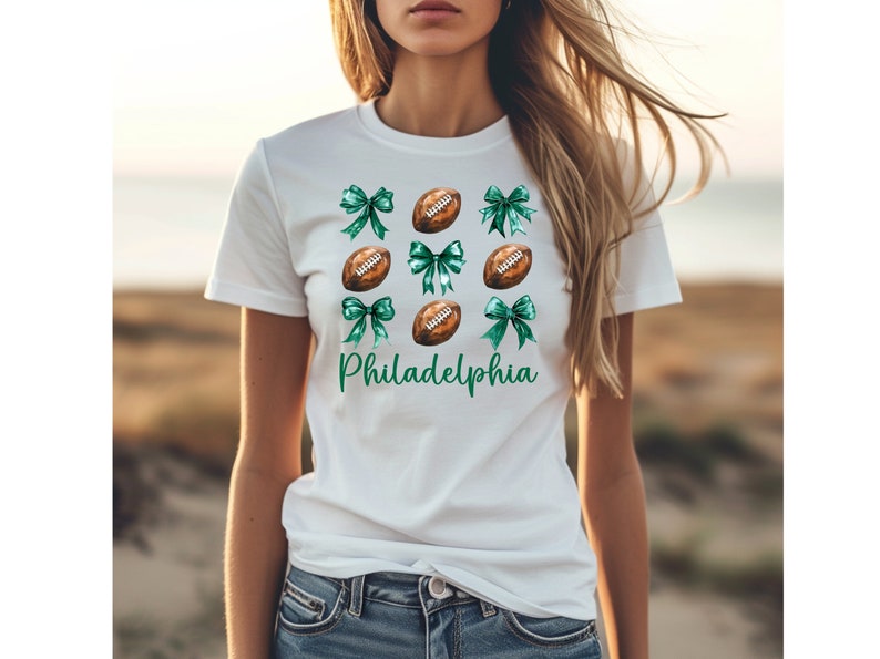 Op de afbeelding: Wit T-shirt met een groen en bruin voetbal- en strikdesign. De tekst "Philadelphia" is op het shirt gedrukt.