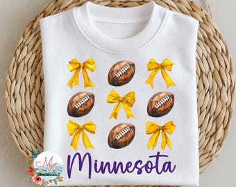 May include: White sweatshirt with a purple "Minnesota" text and a pattern of yellow bows and brown footballs.