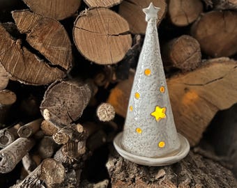 Puede incluir: Un portavelas de cerámica con forma de árbol de Navidad con una estrella en la parte superior. El árbol blanquecino tiene formas de estrella y círculo recortadas que brillan con la luz de la vela en su interior. El portavelas se asienta sobre una pequeña base redonda.