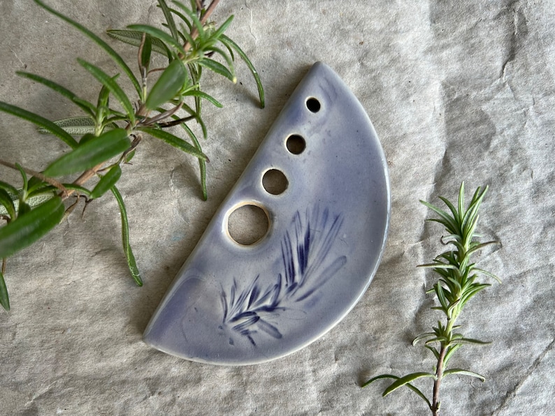 Puede incluir: Un plato de cer&aacute;mica en forma de semic&iacute;rculo con un glaseado lavanda y un dise&ntilde;o de rama de romero. El plato tiene cinco agujeros en una fila a lo largo del borde superior.