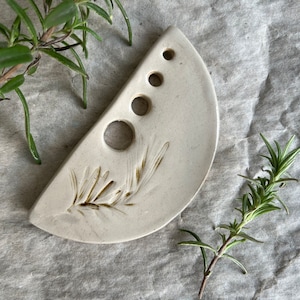 Puede incluir: Un plato de cer&aacute;mica blanca con forma de media luna con cinco agujeros y un dise&ntilde;o de rama de romero. El plato est&aacute; sobre una superficie beige texturizada.