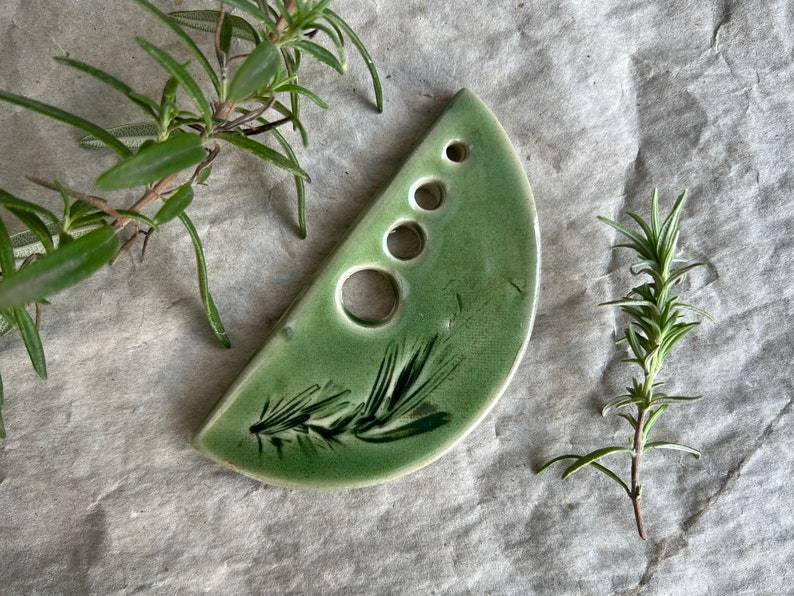 Puede incluir: Un plato de cer&aacute;mica verde en forma de semic&iacute;rculo con cinco agujeros y un dise&ntilde;o de rama de romero. El plato es perfecto para guardar hierbas o especias.