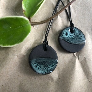 Op de afbeelding: Twee zwarte keramische hangers met een groene en witte glazuur. De hangers liggen op een bruine papieren achtergrond en hangen aan zwarte koorden.