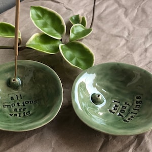 Könnte beinhalten: Zwei grüne Keramik-Räucherstäbchenhalter mit Text. Einer lautet "alle Emotionen sind gültig" und der andere "fühle deine Gefühle". Ein Räucherstäbchen befindet sich in einem Halter. Grüne Blätter sind im Hintergrund.