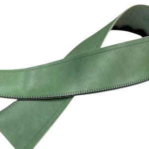 Pode incluir: Uma fita métrica de couro verde com uma borda preta e marcações de medição. A fita é curvada e parece flexível. O fundo é branco.
