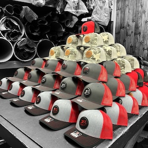 May include: A collection of trucker hats with mesh backs and structured front panels. The hats are in various colours, including red, grey, and camouflage. Each hat has a circular logo patch. The hats are stacked on a grey surface. The image is in monochrome.