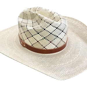 May include: A white straw cowboy hat with a brown leather band and a diamond-shaped pattern. The hat has a wide brim and a slightly pointed crown.