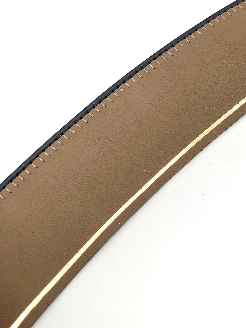 May include: Close-up of a brown leather strap with black stitching and a gold trim. The strap is curved and appears to be part of a larger item, such as a bag or a belt.