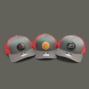May include: Three grey and red baseball caps with a leather patch on the front. The patches feature a stylised letter B with a small design inside.