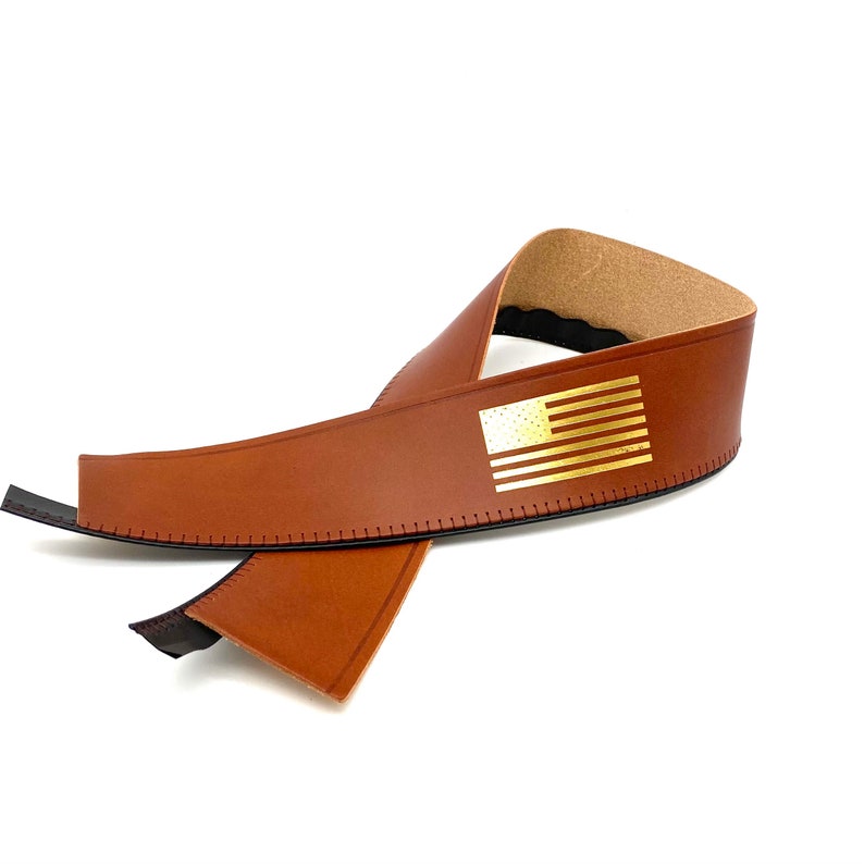 May include: A brown leather guitar strap with a gold American flag design. The strap is folded in half and the flag is visible on the top side.