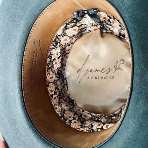 May include: The inside of a gray felt hat with a brown leather band and a floral patterned fabric lining. The lining has a black bow and the text "d.james A FINE HAT CO." printed on it.