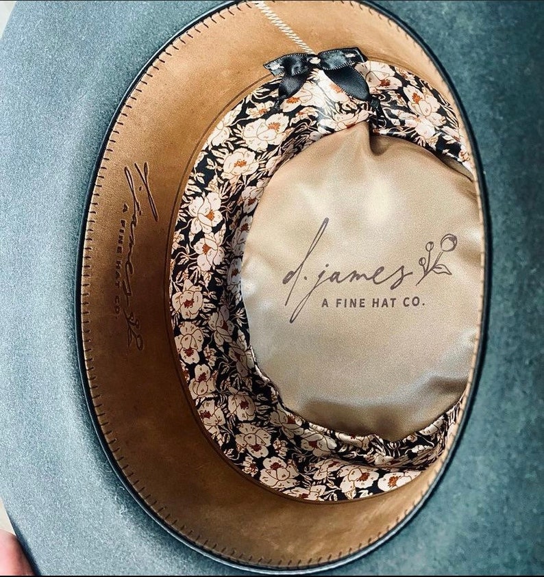 May include: The inside of a gray felt hat with a brown leather band and a floral patterned fabric lining. The lining has a black bow and the text "d.james A FINE HAT CO." printed on it.