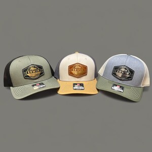 May include: Three trucker hats with a black, beige, and grey mesh back. Each hat has a brown leather patch with gold lettering that reads "Erwin" and a small graphic of a boat.