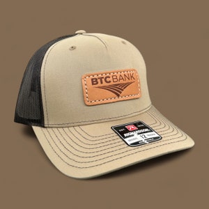 May include: A tan and black trucker hat with a brown leather patch that says "BTCBANK" in brown text. The hat has a black mesh back and a Richardson brand label.