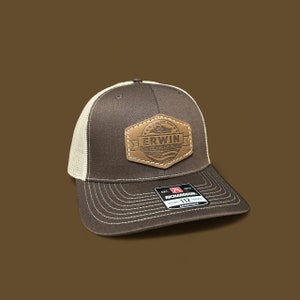 Op de afbeelding: Bruine en beige trucker cap met een bruin leren patch waarop "Erwin Concrete" in witte letters staat. De patch heeft een gestikte rand en een klein beeld van een betonmixer. De pet heeft een zwarte verstelbare band met een wit "Richardson 112" label.