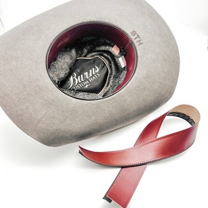 May include: A gray felt cowboy hat with a brown leather band. The hat has a burgundy lining and a black label that says "Burns Custom Hats".