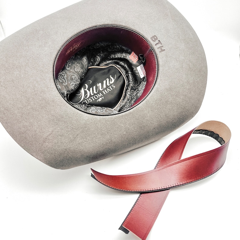 May include: A gray felt cowboy hat with a brown leather band. The hat has a burgundy lining and a black label that says "Burns Custom Hats".