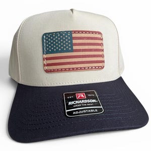 May include: A baseball cap with a light beige crown and a dark navy blue brim. A rectangular patch featuring the United States flag is centered on the front. A black sticker on the brim displays the text: EST. 1970 RICHARDSON WEAR THE BEST ADJUSTABLE.