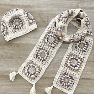 Può includere: Un set di cappello e sciarpa all'uncinetto in beige, marrone e grigio con un motivo geometrico. Il set presenta frange alle estremità della sciarpa.