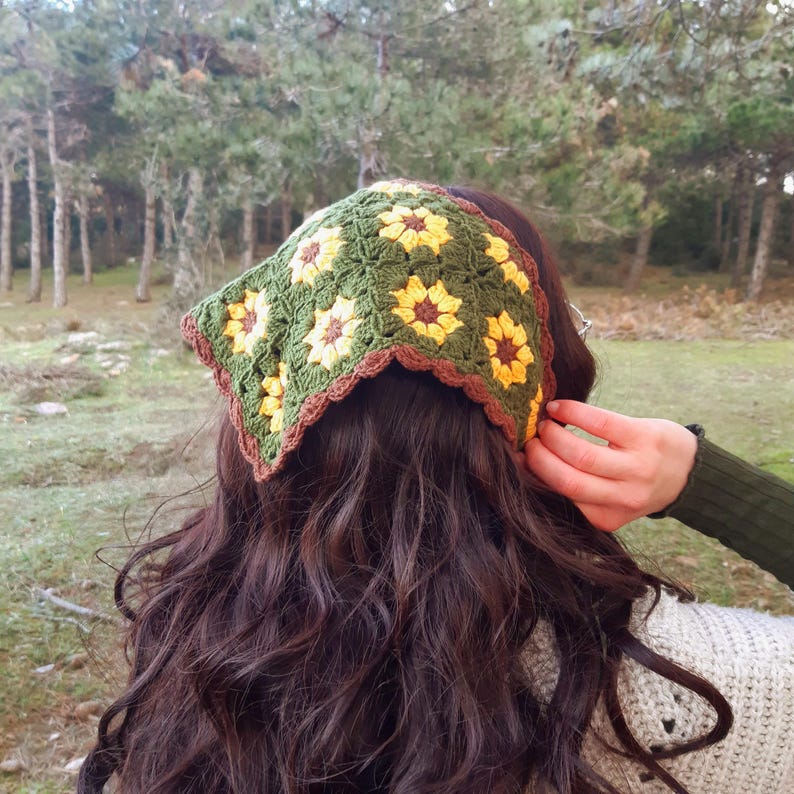 May include: A green crochet headband with yellow and brown flower designs. The headband is square-shaped and has a brown scalloped edge. The person in the image is wearing the headband with their long, dark brown hair.