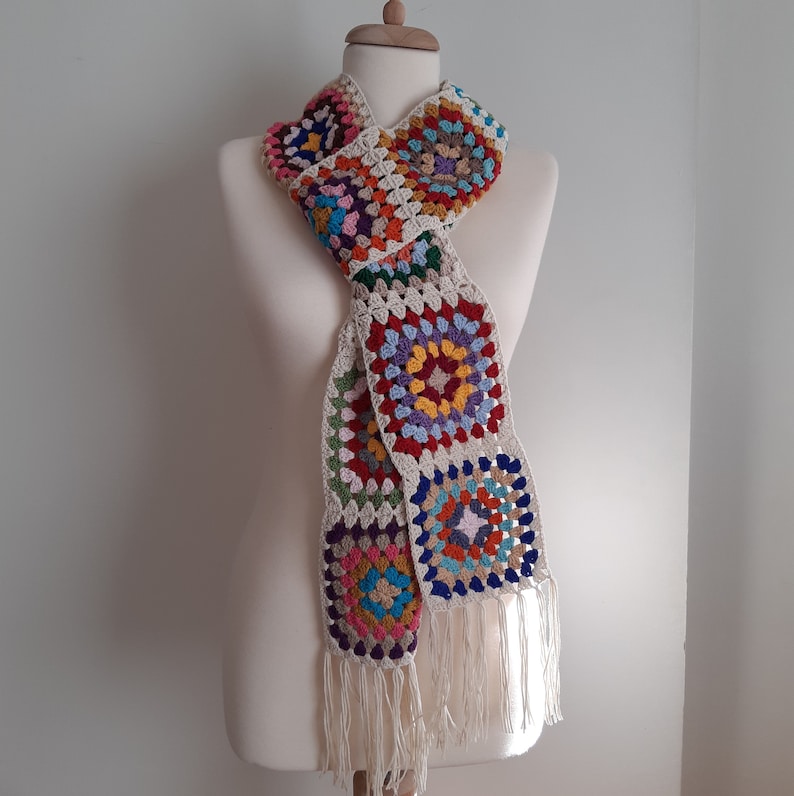 May include: A crocheted scarf with a colorful granny square pattern in shades of red, orange, yellow, green, blue, and purple. The scarf has a fringe at the ends.