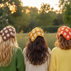 May include: Three women wearing crochet headbands with daisy patterns. The headbands are brown, yellow, and orange, with white daisy accents. The women are facing away from the camera, with long hair and wearing sweaters in green, cream, and yellow.