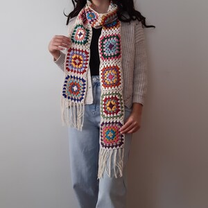 May include: A white crocheted scarf with colorful square patterns. The scarf has fringe on the ends and is worn by a person.