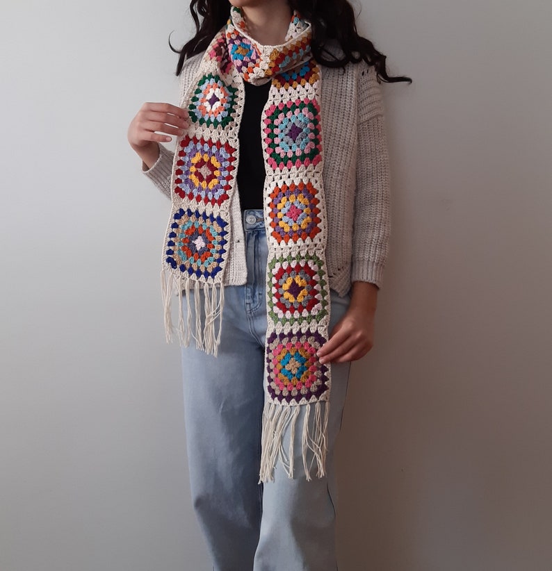 May include: A white crocheted scarf with colorful square patterns. The scarf has fringe on the ends and is worn by a person.