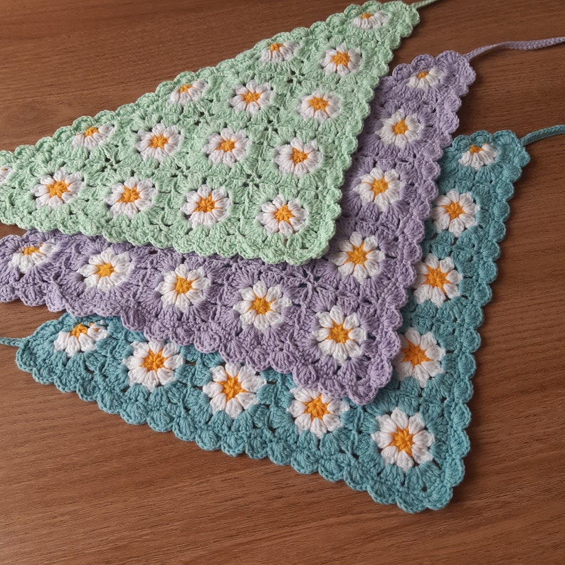 May include: Four crocheted daisy chain banners in pastel colors: light green, lavender, and teal. Each banner is triangular, with white and yellow daisy flowers. The banners are handmade and suitable for home decor.