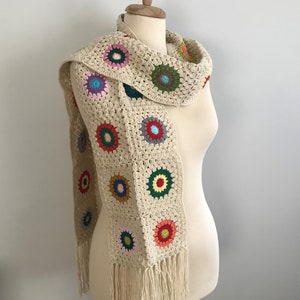 May include: A crocheted scarf with a fringe. The scarf is made of beige yarn and features colorful circular motifs.