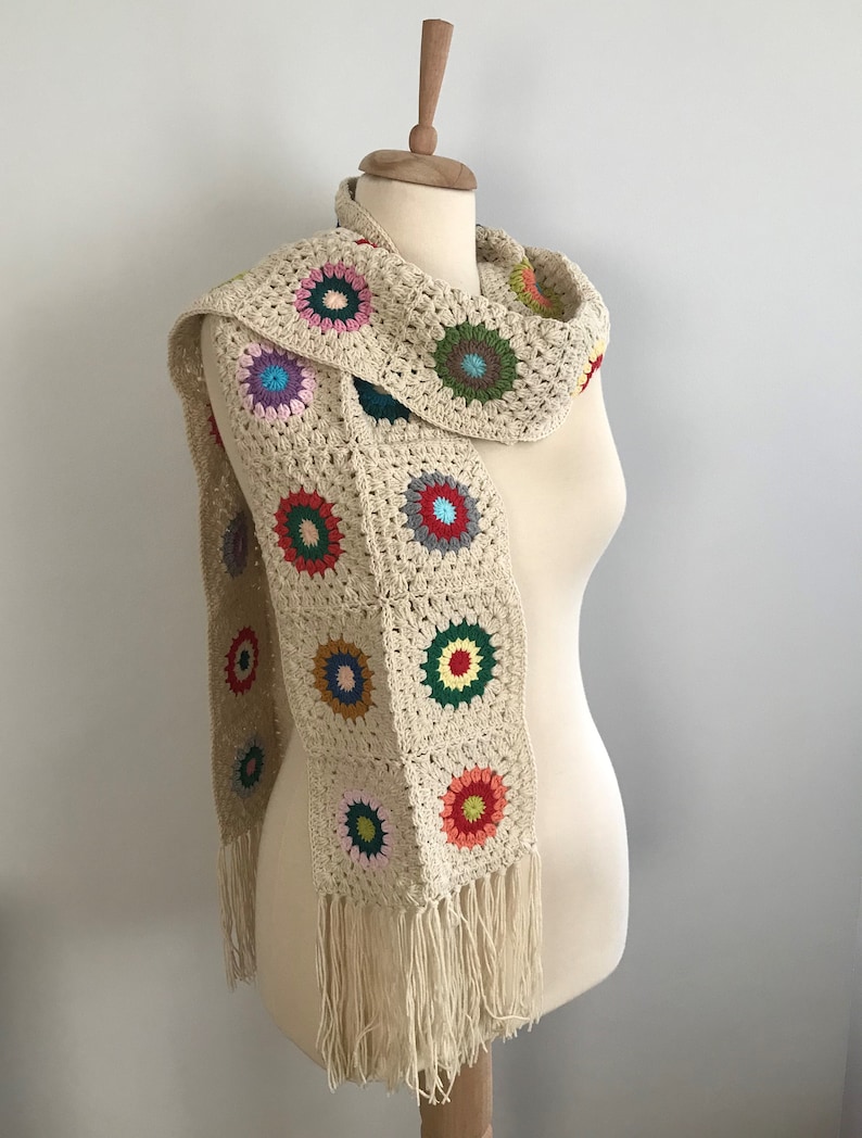May include: A crocheted scarf with a fringe. The scarf is made of beige yarn and features colorful circular motifs.