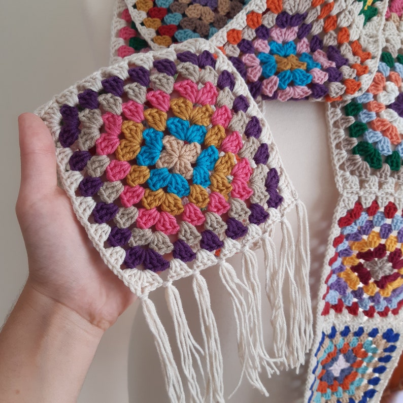 May include: A colorful crocheted scarf with a granny square pattern in shades of pink, purple, blue, yellow, and green. The scarf has a fringe trim and is made with a white yarn.