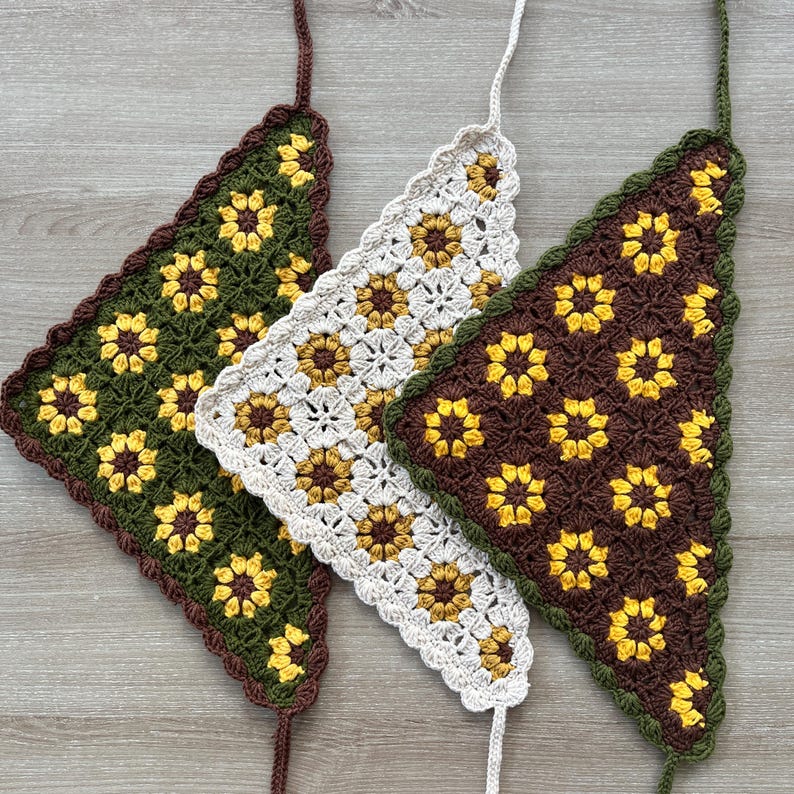 May include: Three crocheted triangle bandanas with sunflower patterns. One is olive green with brown trim, another is off-white with white trim, and the third is brown with green trim. Each has a long tie.