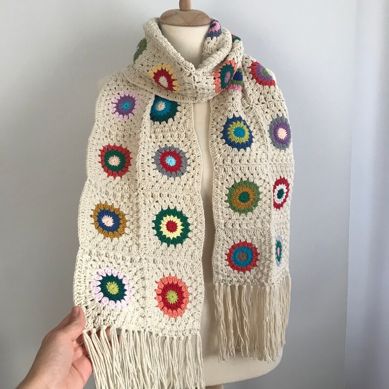 May include: A crocheted scarf with a white background and colorful circular designs. The scarf has fringe on the ends.