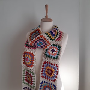 May include: A colorful crocheted scarf with a granny square pattern in shades of red, orange, yellow, green, blue, and purple. The scarf is made with a cream-colored yarn.