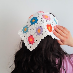 May include: A white crochet headband featuring colorful floral designs in blue, pink, orange, and purple. The headband is handmade with a scalloped edge, offering a vibrant and stylish accessory.