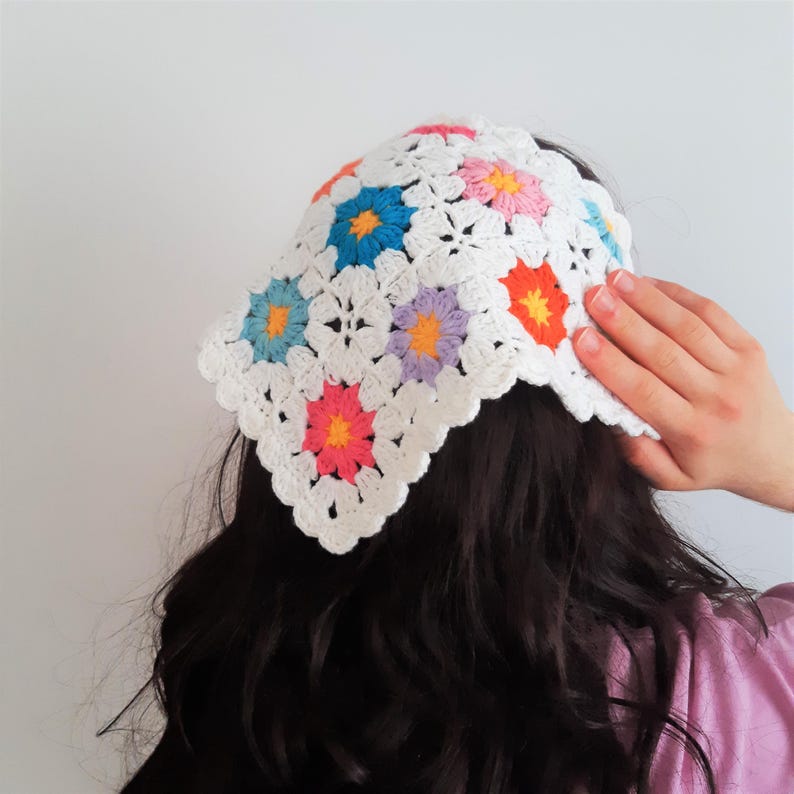 May include: A white crochet headband featuring colorful floral designs in blue, pink, orange, and purple. The headband is handmade with a scalloped edge, offering a vibrant and stylish accessory.