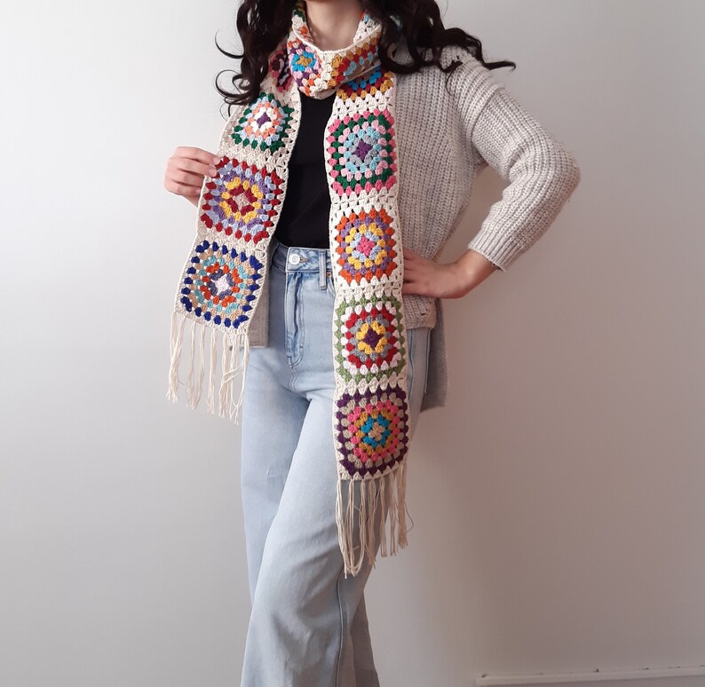May include: A colorful crocheted scarf with a fringe. The scarf is made of white yarn with multi-colored squares. The scarf is worn by a person wearing a white sweater and blue jeans.